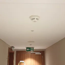 Korytarz z wykończonym sufitem podwieszanym, widoczne czujniki dymu i oświetlenie, ściany w kolorze beżowym, drzwi z oznaczeniem drogi ewakuacyjnej.