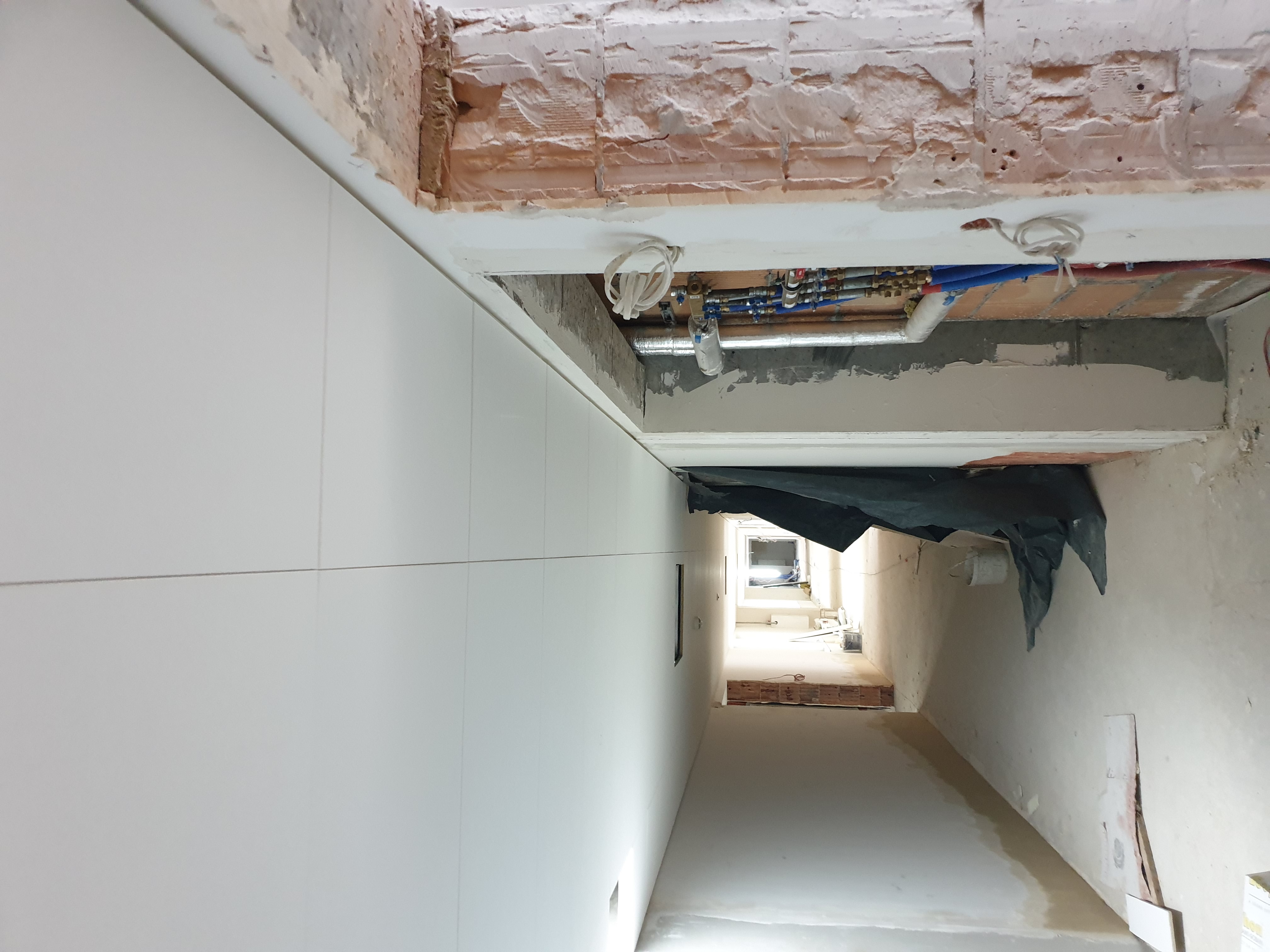 Wnętrze w trakcie remontu: widoczne instalacje wodne pod sufitem, surowy mur ceglany, białe płyty na ścianie i zabezpieczenie folią malarską.