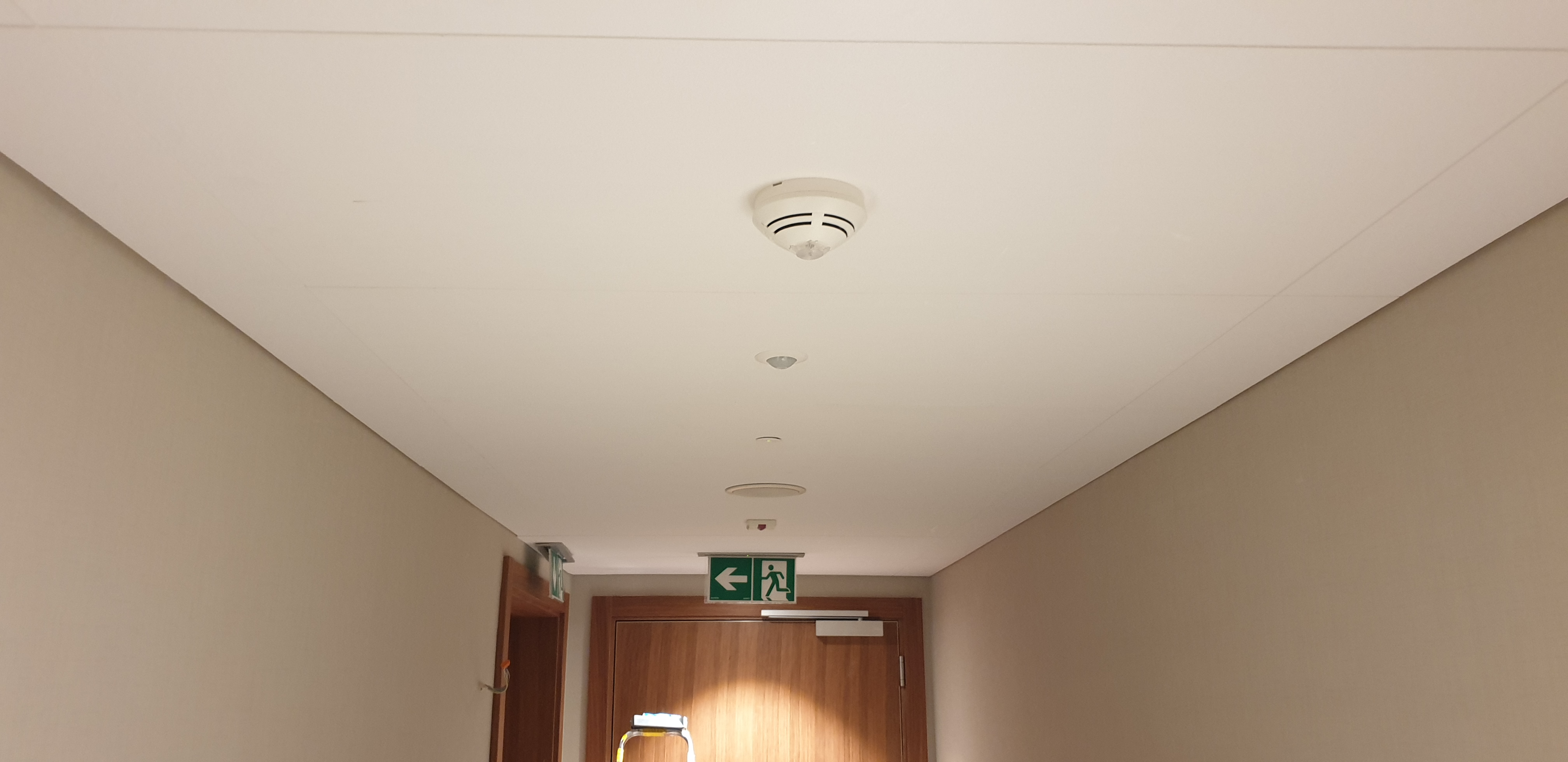 Korytarz z wykończonym sufitem podwieszanym, widoczne czujniki dymu i oświetlenie, ściany w kolorze beżowym, drzwi z oznaczeniem drogi ewakuacyjnej.