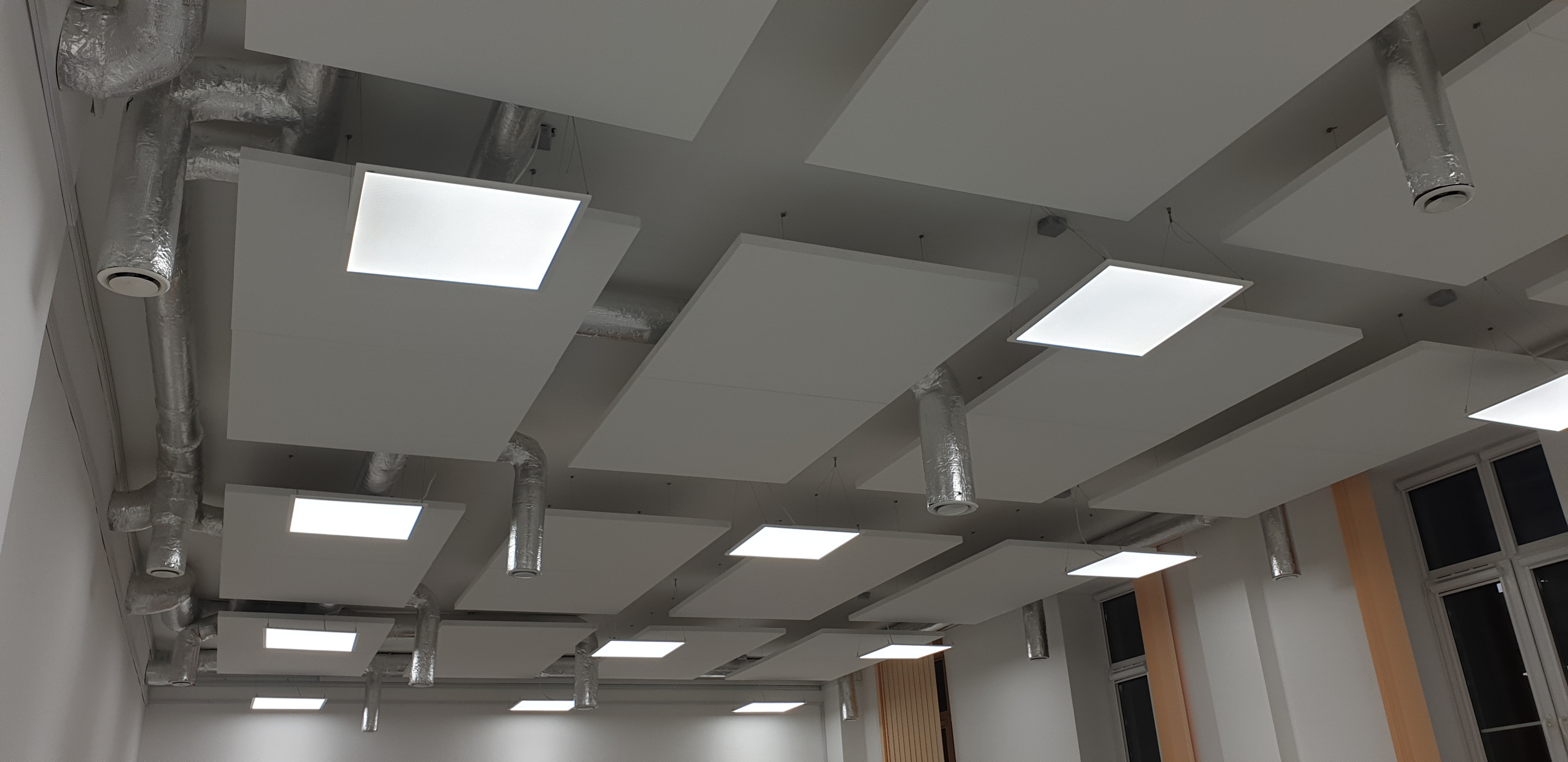 Sufit podwieszany z kwadratowymi panelami i oświetleniem LED, widoczne instalacje wentylacyjne w srebrnej izolacji.