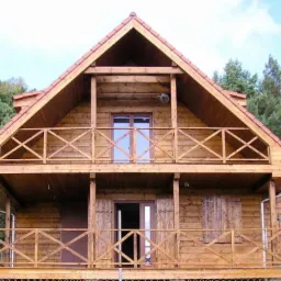 Dwukondygnacyjny dom z drewna z balkonami, widoczny na tle zieleni i błękitnego nieba. Czerwona dachówka. Charakterystyczne balustrady balkonowe w kształcie krzyży.