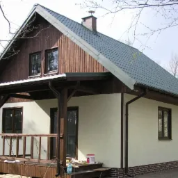 Ukończony dom z drewnianą elewacją na poddaszu i jasnymi ścianami. Widoczny drewniany taras z balustradą i fragment dachu z kominem. Detal: farba na schodach.