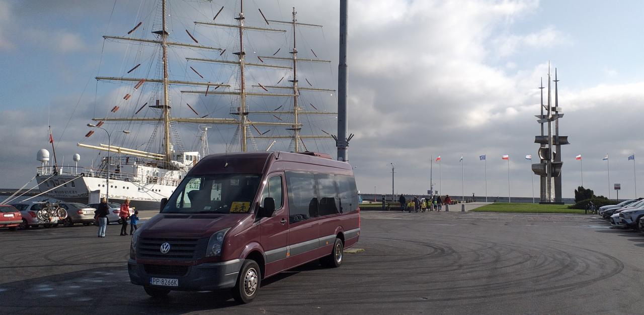 Ciemnoczerwony bus Volkswagen Crafter z żółtą naklejką przewozu osób niepełnosprawnych na przedniej szybie, zaparkowany na parkingu w Gdyni z widokiem na Dar Młodzieży i pomnik Polski Morskiej