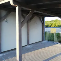 Zadaszona przestrzeń z drewnianą konstrukcją wspartą na filarach, z widocznymi elementami montażowymi i surowym betonowym podłożem, otoczona metalową barierką z widokiem na zieleń.