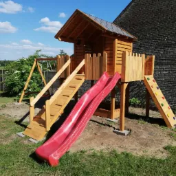 Drewniany plac zabaw dla dzieci z domkiem, zjeżdżalnią i ścianką wspinaczkową, stojący na trawniku przed ciemnym budynkiem z cegły.