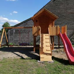 Dremeno - Drewniany plac zabaw z domkiem, schodami, zjeżdżalnią i huśtawką na tle ceglanego budynku w słoneczny dzień.