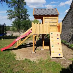 Dremeno - Drewniany plac zabaw z czerwoną zjeżdżalnią i ścianką wspinaczkową z kolorowymi uchwytami, na tle zielonego trawnika i murowanego budynku.