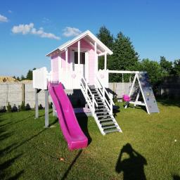 Dremeno - Różowy domek zabaw dla dzieci z białą stolarką, schodami, zjeżdżalnią i ścianką wspinaczkową, ustawiony na trawniku w ogrodzie.