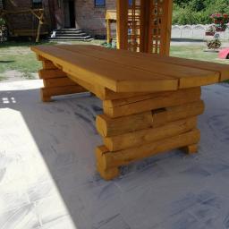 Dremeno - Stół ogrodowy wykonany z okrąglaków, z widoczną konstrukcją z bali i grubym blatem, ustawiony na jasnej, wyłożonej płytkami powierzchni, z fragmentem altany w tle.