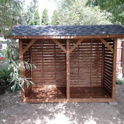 Dremeno - Drewniana konstrukcja z zadaszeniem z gontu bitumicznego, podzielona na dwie części, z poziomymi listwami, umieszczona w ogrodzie z widocznym drzewem i ogrodzeniem z cegły.