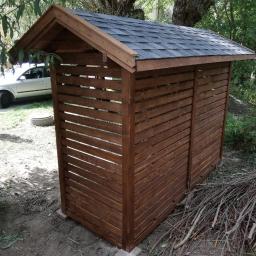 Dremeno - Drewniana konstrukcja z dachem krytym gontem bitumicznym, prawdopodobnie drewutnia, z poziomymi szczelinami, stojąca na zewnątrz w otoczeniu drzew i gałęzi.