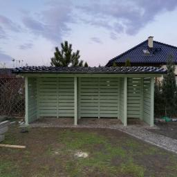 Dremeno - Nowo wybudowana, jasna, drewniana konstrukcja z zadaszeniem z dachówki, podzielona na trzy otwarte przestrzenie, na tle domu z ciemną dachówką i zielenią.