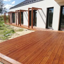 Nowoczesny dom z jasną elewacją i rozległym tarasem z desek kompozytowych, drewniane słupki podtrzymujące konstrukcję przy ścianie budynku, widok na ogród.