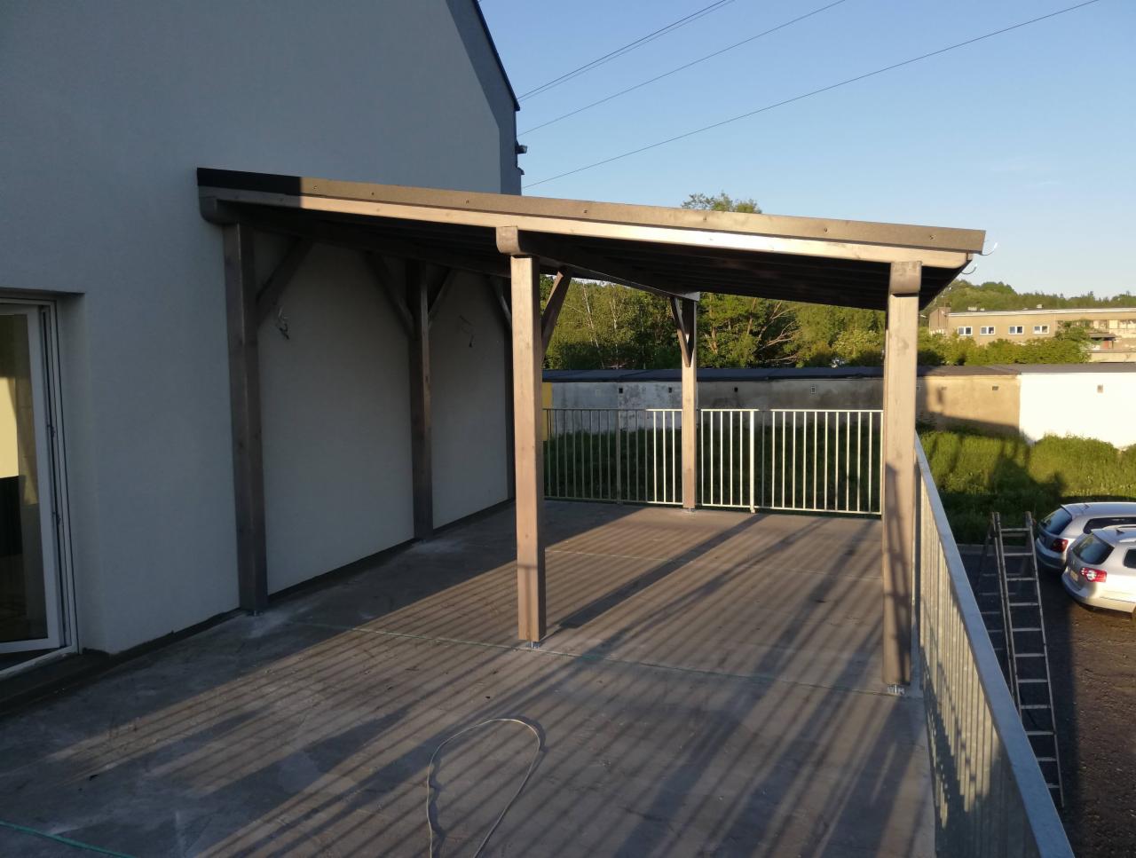 Zadaszony taras przylegający do budynku, z drewnianą konstrukcją wspartą na słupach, widok na metalową barierkę, drabina oparta o samochód w tle.