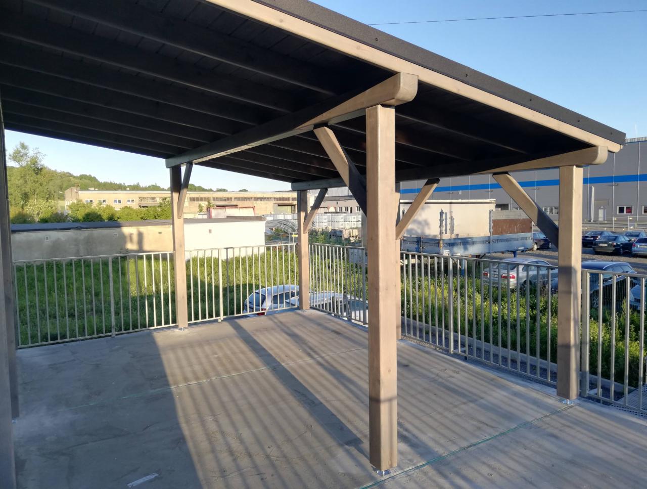 Zadaszony obszar z drewnianymi słupami i metalowymi barierkami, widok na parking i budynki w tle.