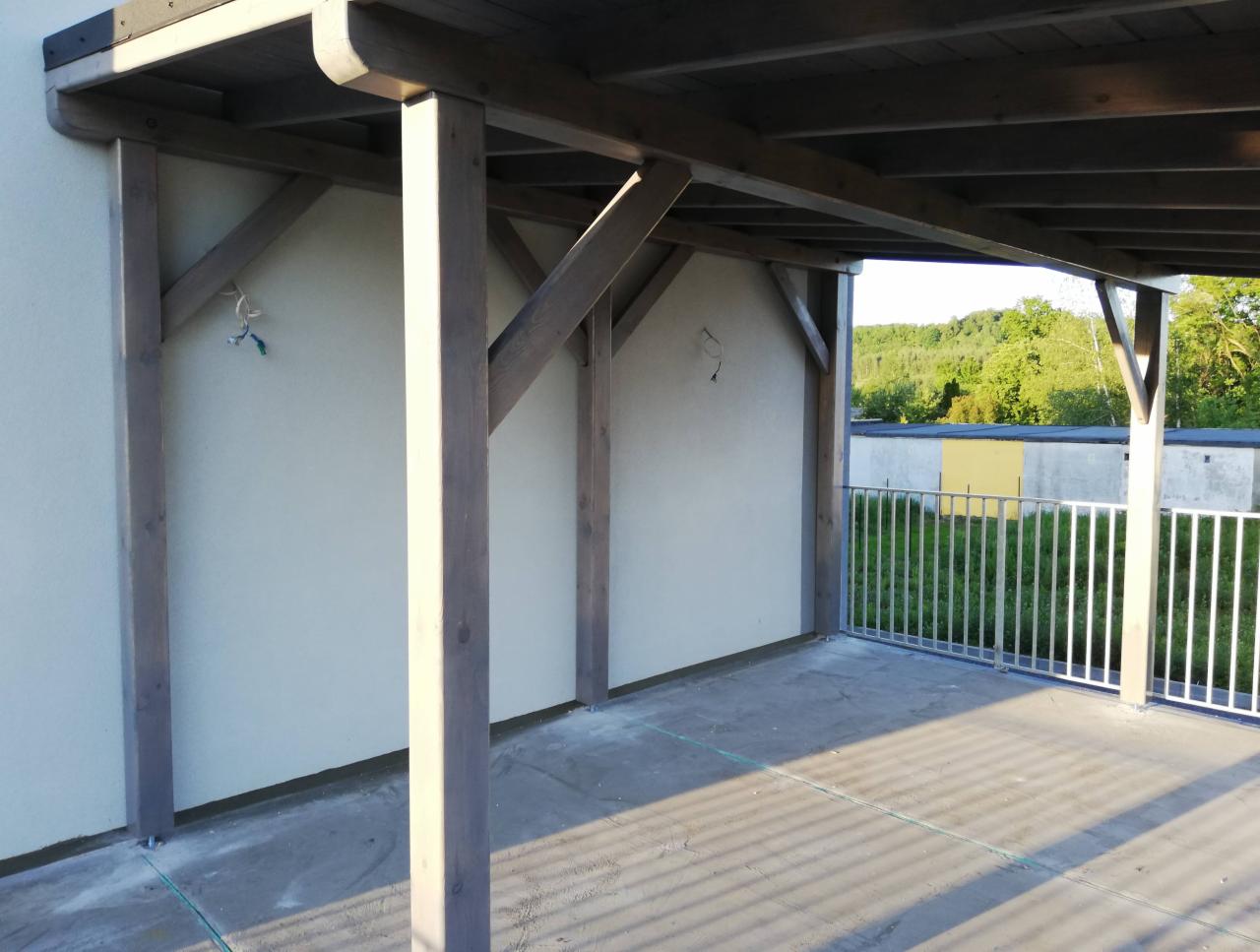 Zadaszona przestrzeń z drewnianą konstrukcją wspartą na filarach, z widocznymi elementami montażowymi i surowym betonowym podłożem, otoczona metalową barierką z widokiem na zieleń.