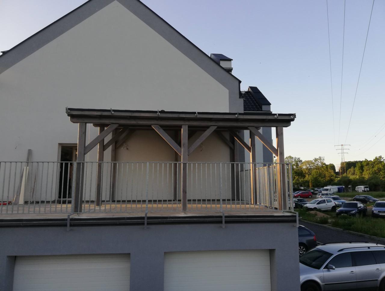 Drewniana konstrukcja zadaszenia na tarasie z metalową balustradą, widok z boku na tle parkingu i linii wysokiego napięcia.