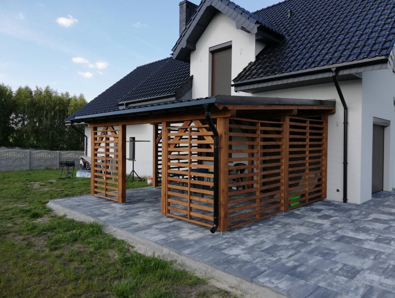 Drewniana konstrukcja z poziomymi listwami, przylegająca do białego domu z ciemnym dachem, tworząca zadaszone miejsce na szarym brukowanym podłożu. Widoczne rynny i elementy mocujące do budynku.