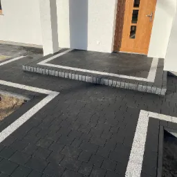 Ułożona czarna kostka brukowa z jasnymi obrzeżami prowadząca do drzwi wejściowych domu. Widoczne schody z kostki.