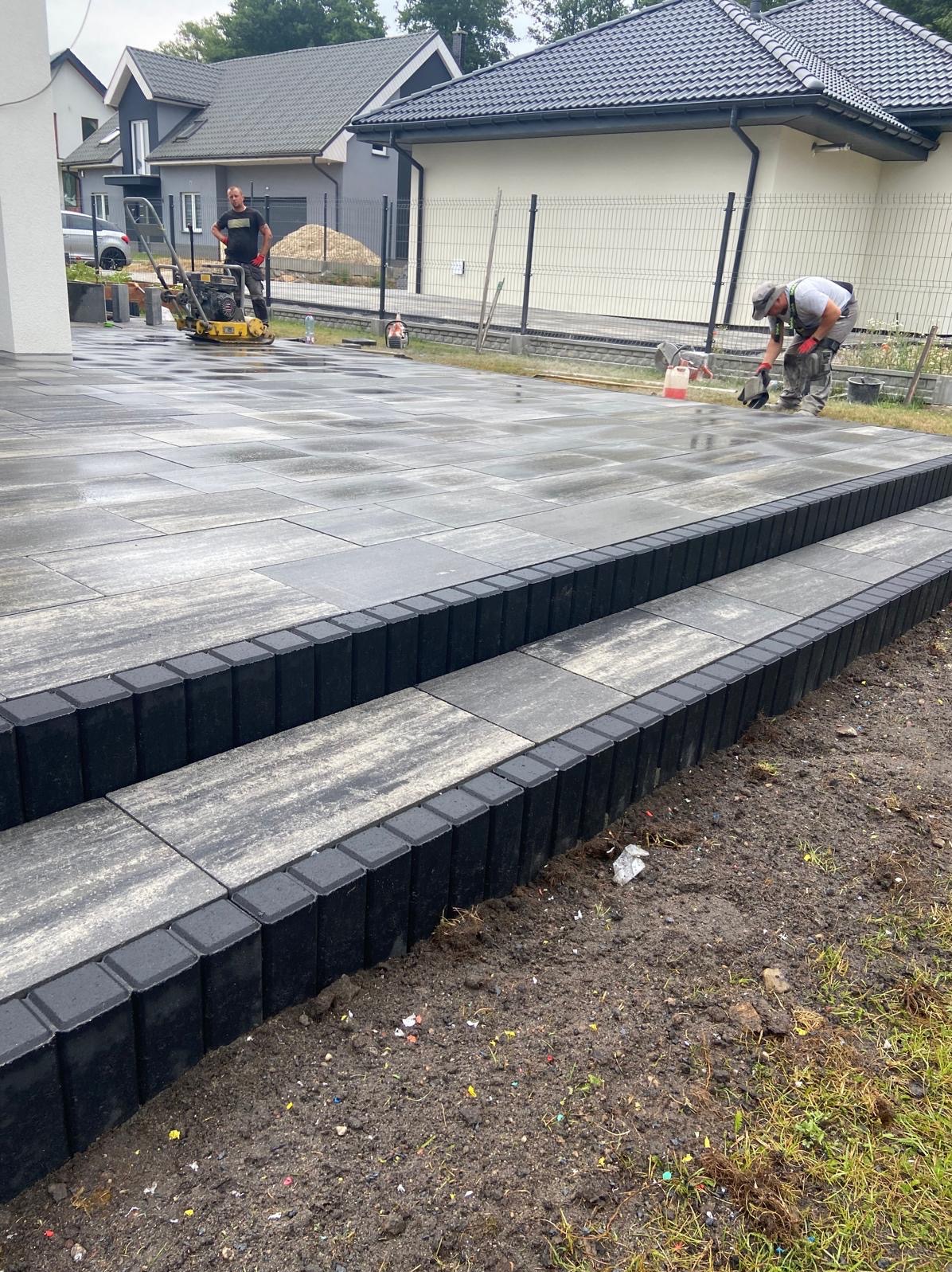 Ułożona kostka brukowa tworząca schody wejściowe. W tle pracownicy i maszyna do ubijania. Nowoczesny dom z szarą elewacją i grafitowym dachem.
