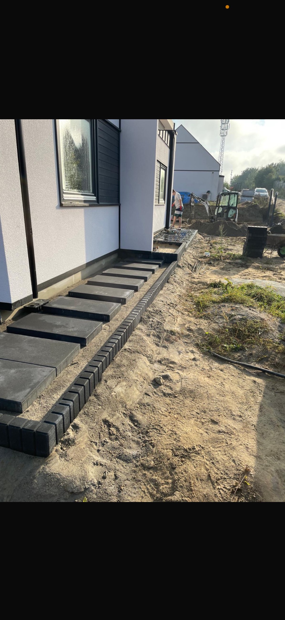 Nowoczesne schody z kostki brukowej prowadzące do domu, obok widoczny wykop i maszyna budowlana. Minimalistyczny design, szara kostka, surowe otoczenie.