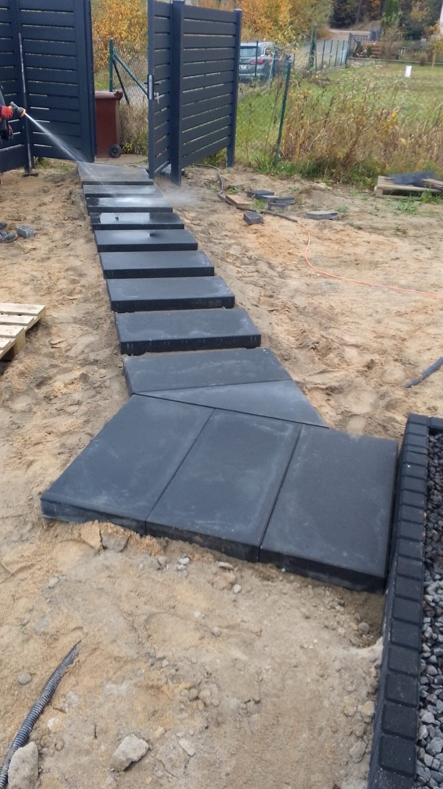 Ścieżka z ciemnych płyt prowadząca do nowoczesnego, grafitowego ogrodzenia panelowego. Płyty są myte wodą pod ciśnieniem. Widoczne elementy otoczenia działki.