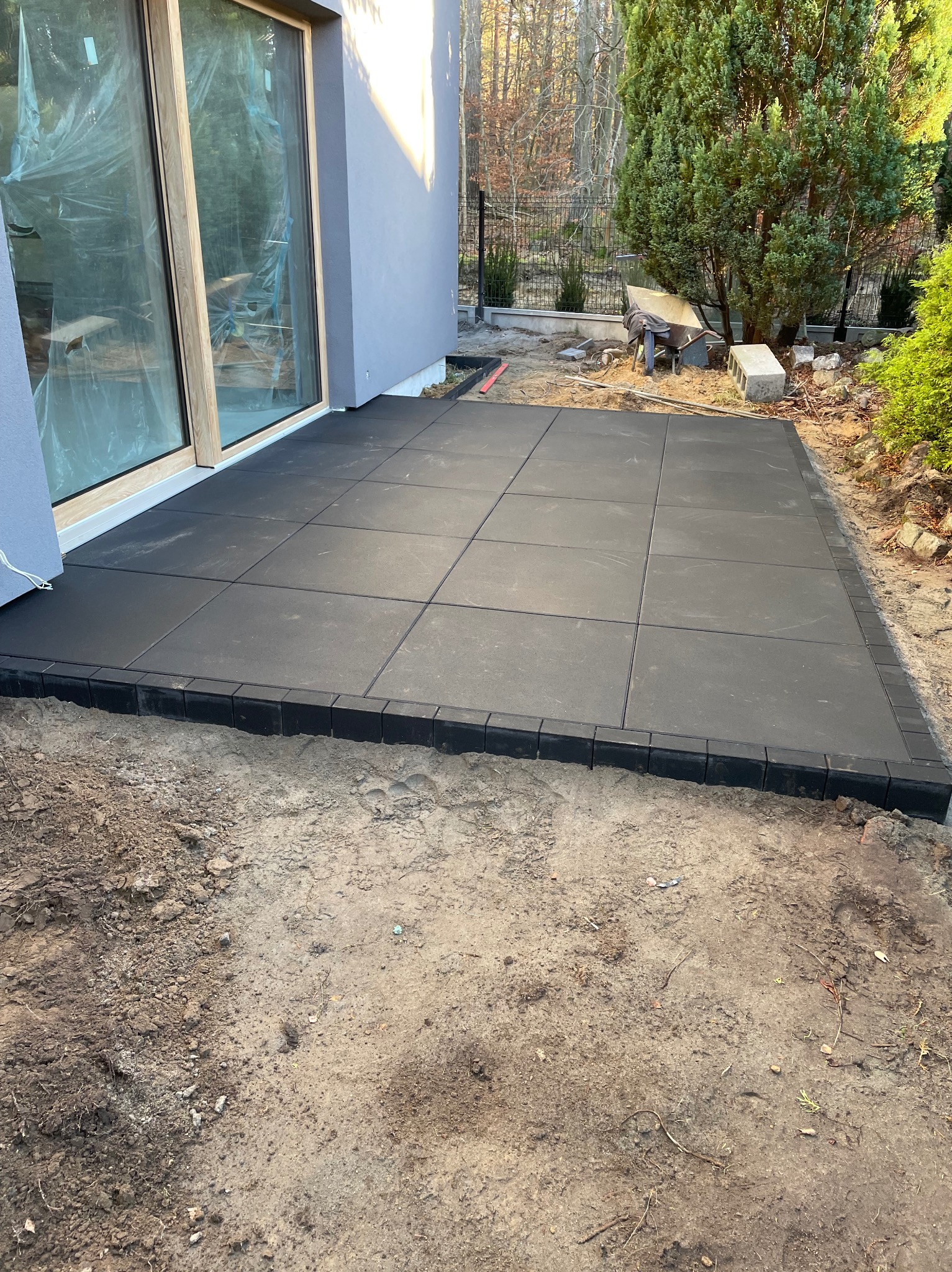 Nowoczesny taras z ciemnoszarej kostki brukowej, obrzeża z kostki, widok na przesuwne drzwi tarasowe i ogród. Minimalistyczny styl, świeżo po realizacji.