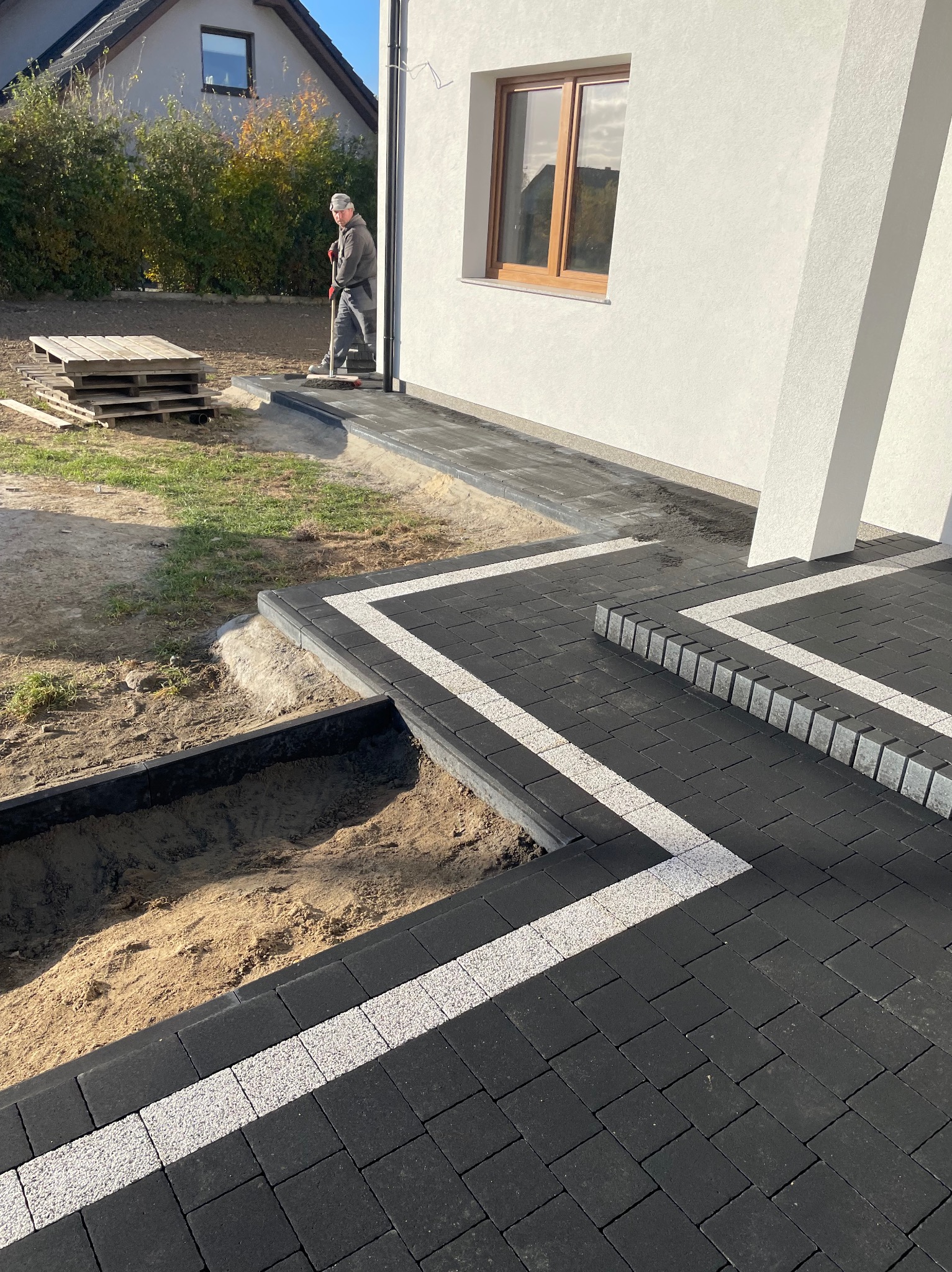 Układanie czarno-białej kostki brukowej na posesji, widoczny pracownik z miotłą. Częściowo wykopany teren i palety z kostką w tle. Nowoczesne wykończenie.