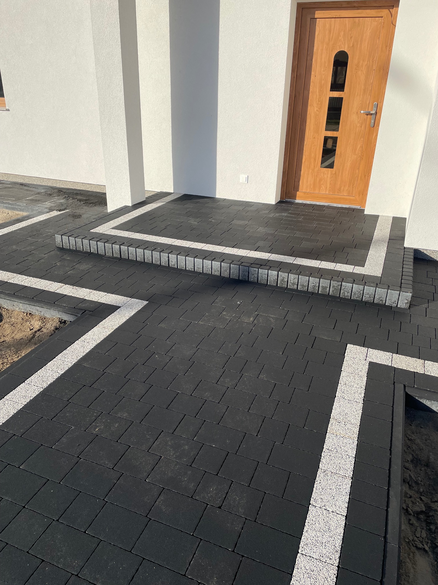 Ułożona czarna kostka brukowa z jasnymi obrzeżami prowadząca do drzwi wejściowych domu. Widoczne schody z kostki.