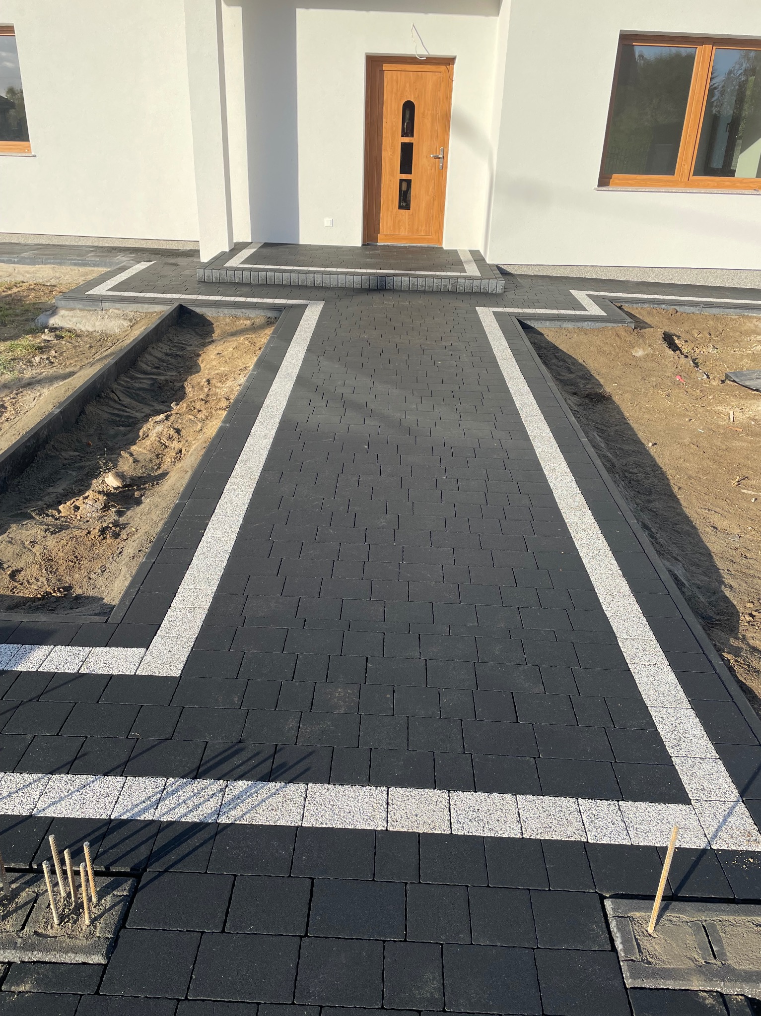 Ułożona czarno-biała kostka brukowa prowadząca do domu w Borkowie. Widoczne obrzeża i fragment wykopu. Nowoczesny design i precyzja wykonania.