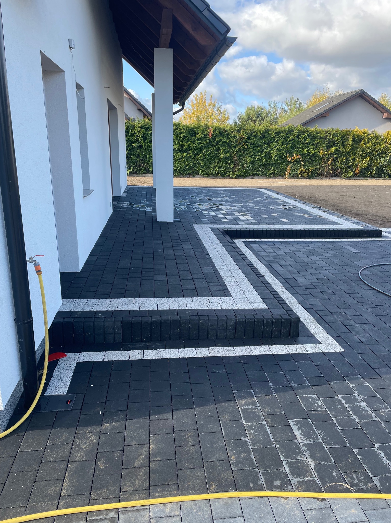 Nowoczesna nawierzchnia z ciemnej kostki brukowej z jasnymi akcentami wokół domu. Widoczne schody z kostki i fragment budynku z rynną.