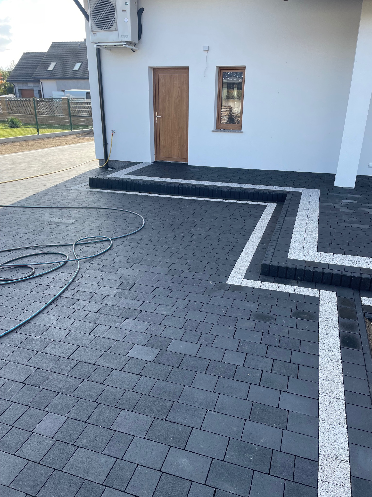 Nowoczesna, grafitowa kostka brukowa z białymi akcentami, tworząca geometryczny wzór przed domem. Wąż ogrodowy leży na nawierzchni.