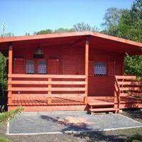 Czerwony, drewniany domek letniskowy z werandą i okiennicami na tle zieleni drzew.