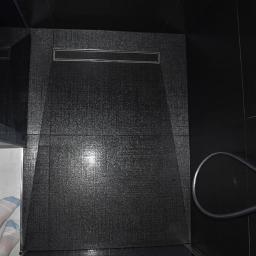 New Home KT Usługi remontowo-wykończeniowe - Czarny brodzik z odpływem liniowym, widoczne stopy w skarpetkach w rogu.