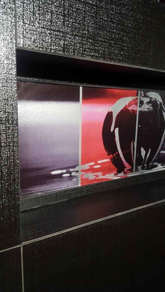 Czarna ściana łazienki z błyszczących płytek z wnęką zawierającą abstrakcyjny, trójdzielny obraz z motywem jabłka z polewą.