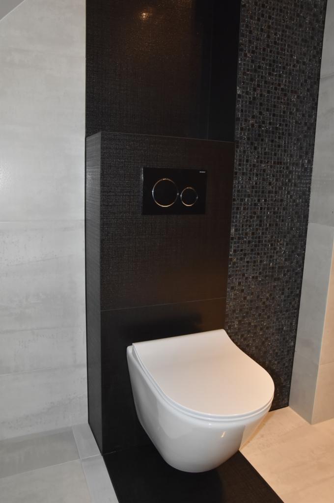 Podwieszana miska WC z czarnym panelem spłukującym na tle mozaiki i jasnych płytek w wykończonej łazience.