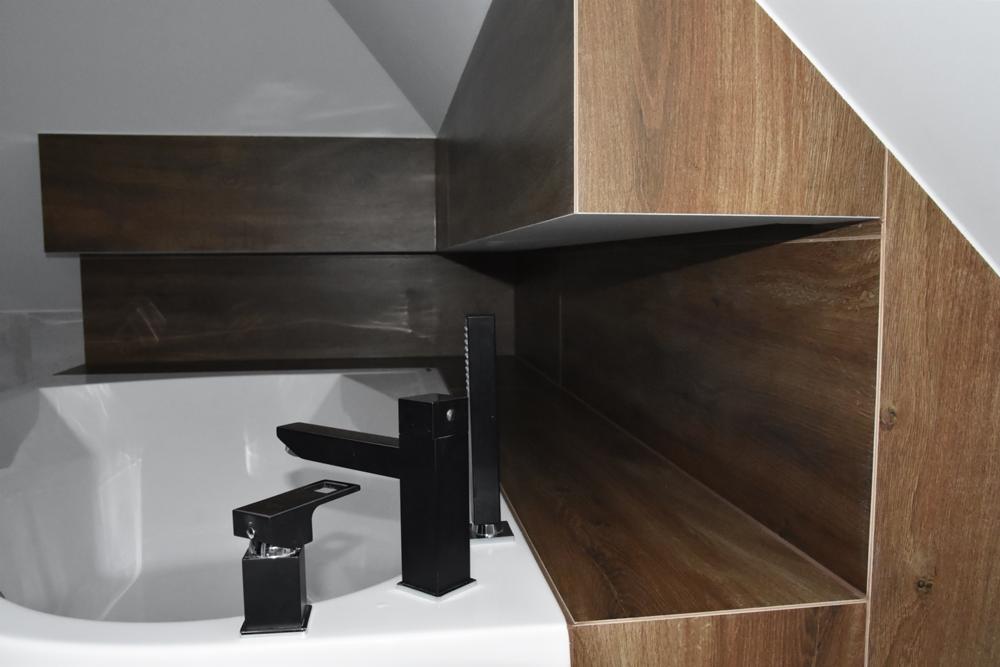 Nowoczesna łazienka na poddaszu z drewnopodobnymi płytkami, białą wanną i czarną, minimalistyczną armaturą.