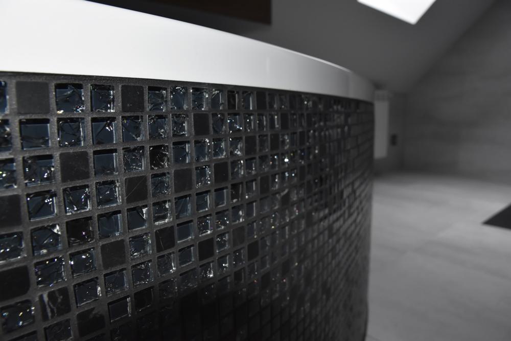 Częściowe ujęcie zaokrąglonej obudowy wanny wyłożonej czarną mozaiką z błyszczącymi elementami w jasnej łazience na poddaszu.