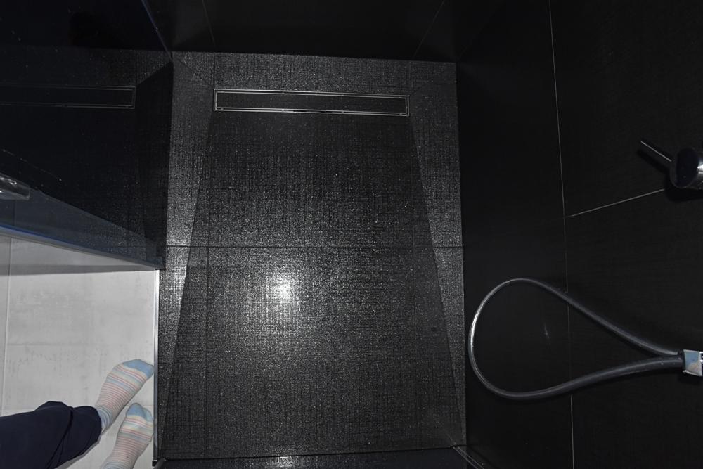 Czarny brodzik z odpływem liniowym, widoczne stopy w skarpetkach w rogu.