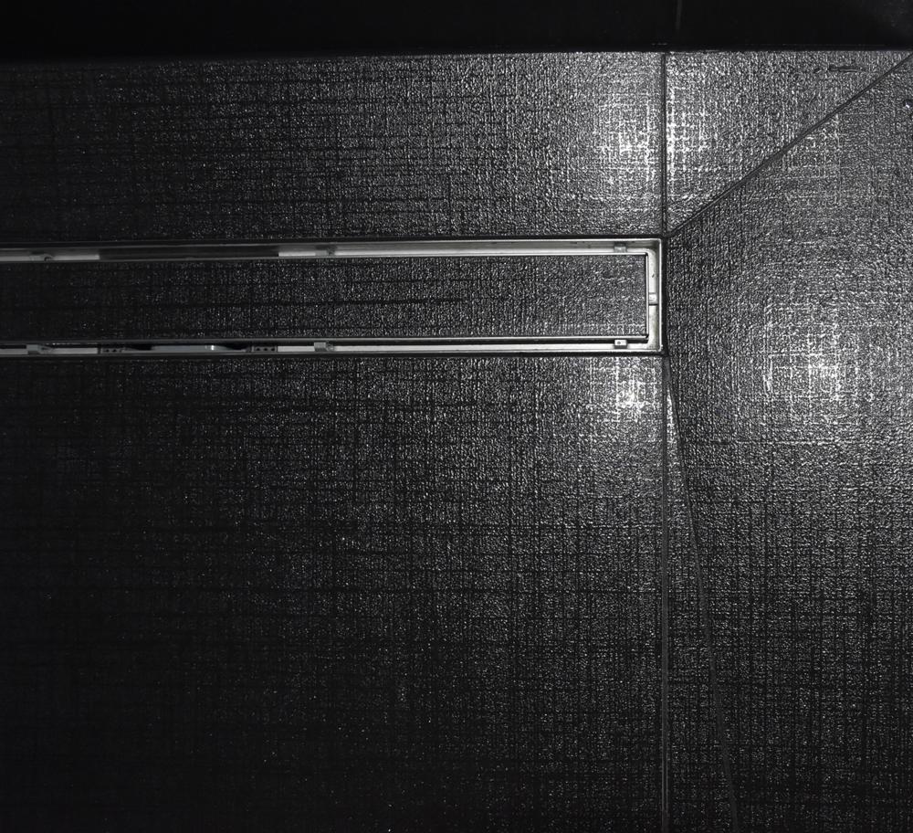 Czarne, teksturowane płytki podłogowe z odpływem liniowym w rogu kabiny prysznicowej, widoczne fugi.