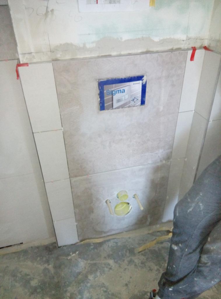 Montaż stelaża podtynkowego Geberit Sigma z widocznymi otworami na przyłącza i częściowo ułożonymi płytkami w trakcie prac wykończeniowych łazienki.