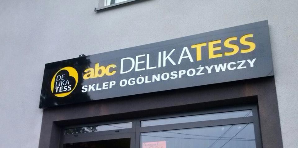 Szyld sklepu spożywczego 'abc Delikatess' z logo i napisem 'Sklep Ogólnospożywczy' na szarym tle, zamontowany nad wejściem do sklepu.