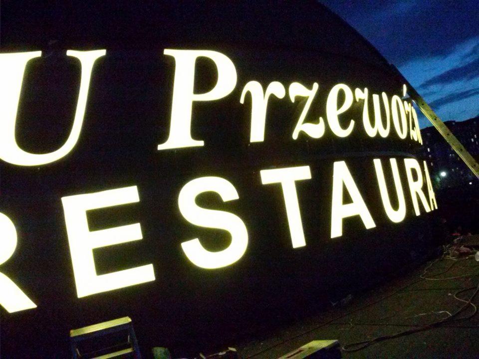 Podświetlany szyld restauracji 'U Przewoźnika' z widocznymi elementami montażowymi i przewodami, sfotografowany o zmierzchu.