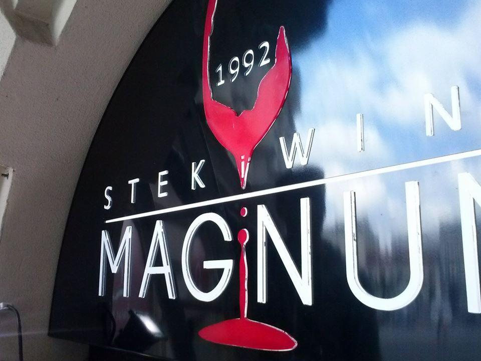 Czarno-czerwony szyld z nazwą 'STEK MAGNUM WIN', rocznikiem 1992 i stylizowanym kieliszkiem czerwonego wina, zamontowany na ścianie.