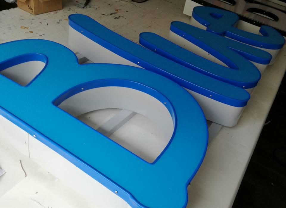 Niebieskie, przestrzenne litery z podświetleniem, wykonane z tworzywa sztucznego, leżące na stole w warsztacie.