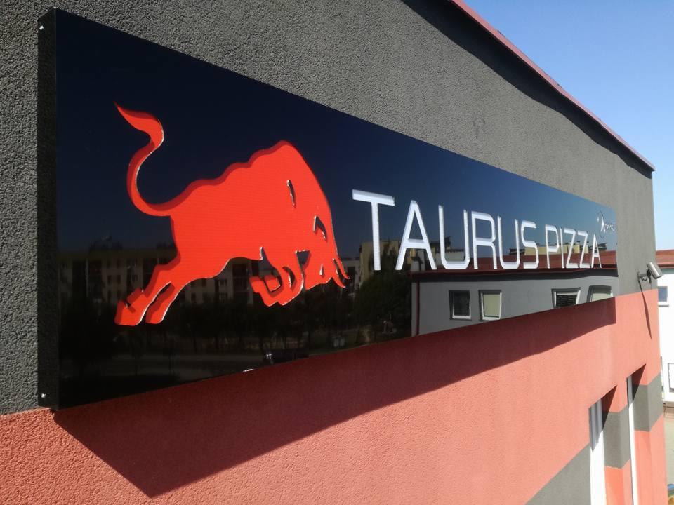 Czarny szyld reklamowy Taurus Pizza z czerwonym logo byka i białym napisem, zamontowany na szaro-pomarańczowej ścianie budynku.
