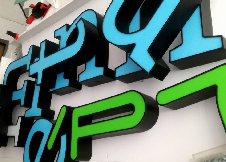 Trójwymiarowe litery w kolorach niebieskim i zielonym z czarnymi obramowaniami, przymocowane do białej ściany, widoczne pod kątem.