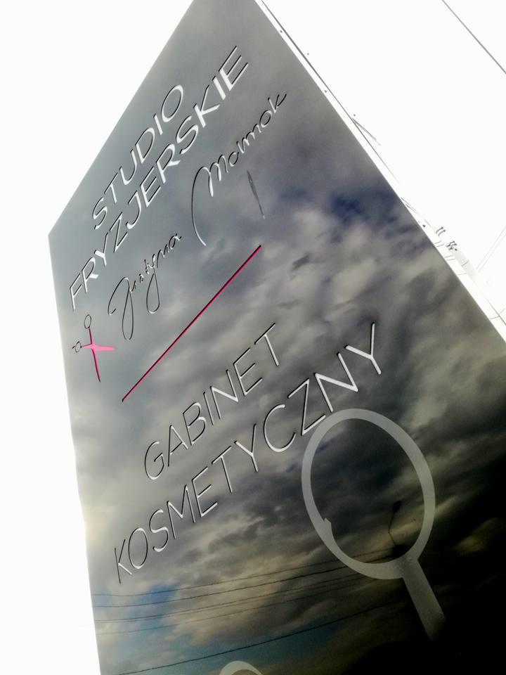 Pylon reklamowy z nazwą studia fryzjerskiego i gabinetu kosmetycznego, wycięte litery na tle pochmurnego nieba, widoczne linie energetyczne w tle.