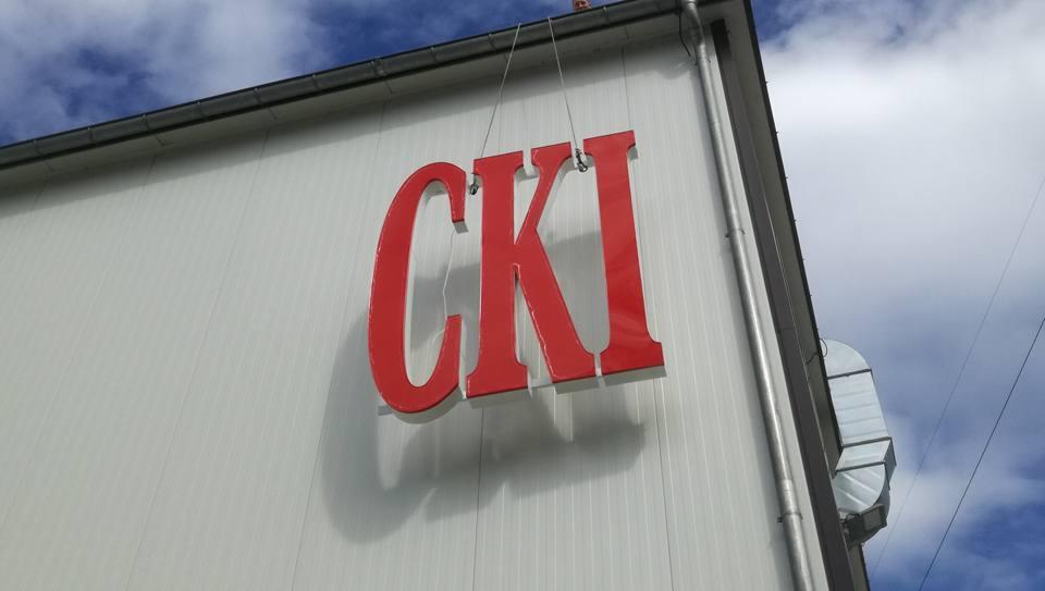 Czerwone litery CKI zamontowane na białej ścianie budynku, widoczne z dołu pod kątem. Litery trójwymiarowe, mocowane na linkach. W tle fragment nieba z chmurami.