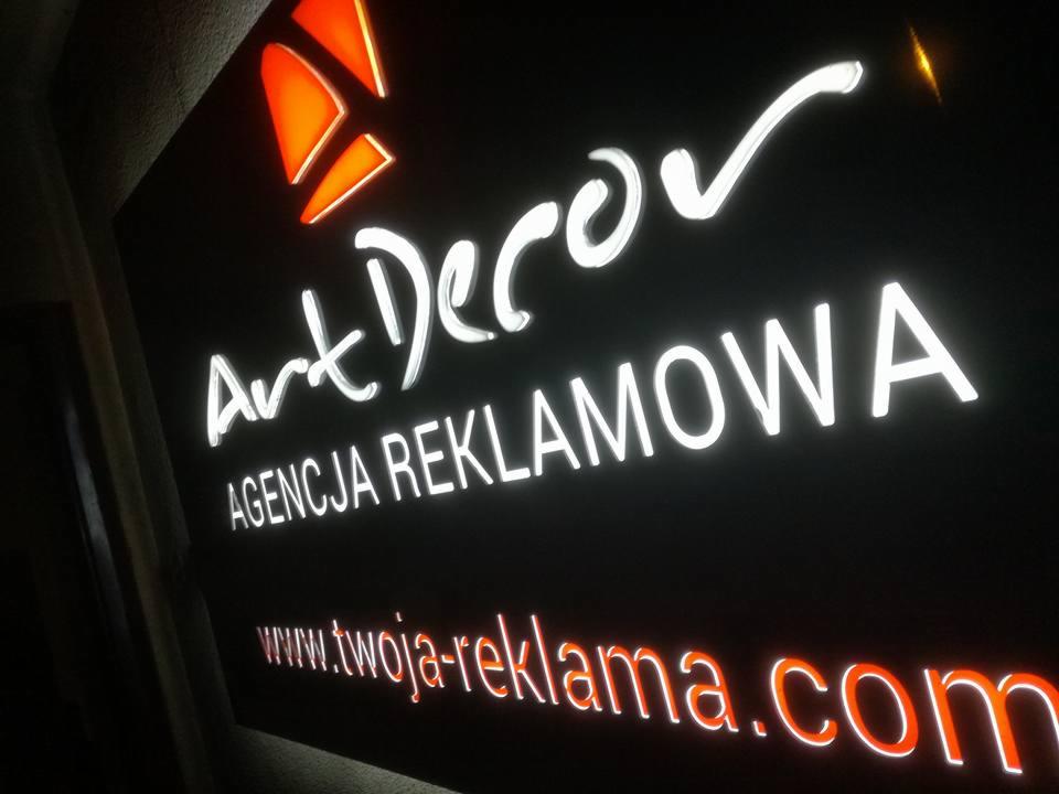 Świecący szyld reklamowy firmy Art Decor z adresem strony internetowej, widoczny pod kątem z dołu.